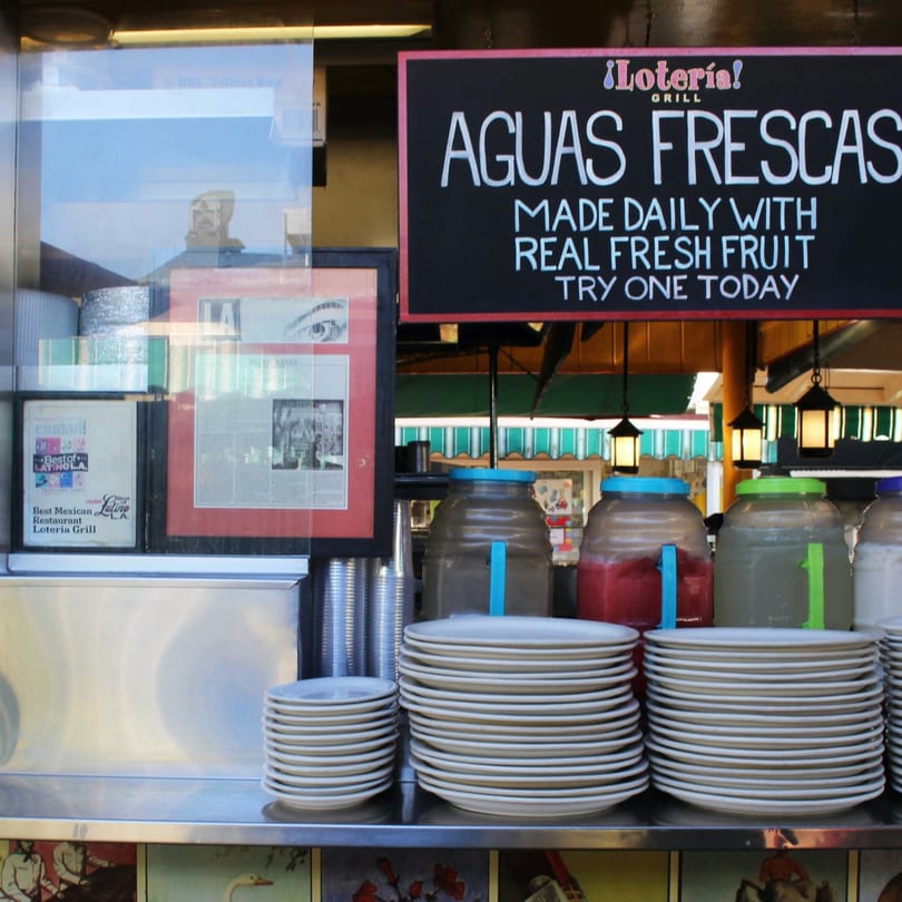 Loteria Grill's Aguas Frescas sign, with large drink dispensers and stacks of plates below.